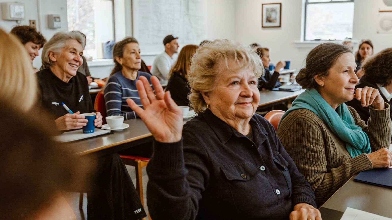 Adultos mayores sentados en una sala comunitaria luminosa levantando la mano para hacer preguntas en una presentación educativa de Medicare